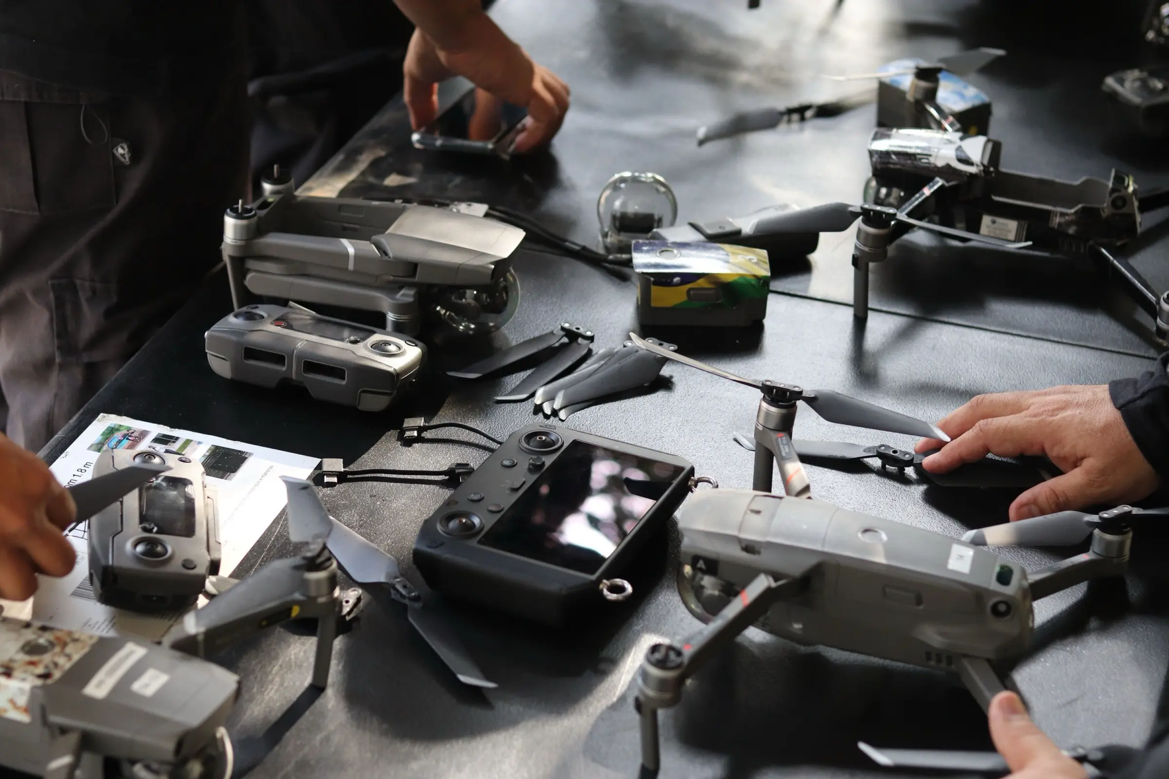 Folding quadcopters, controllers and spare propellers laid out on a dark workbench, with hands reaching in.