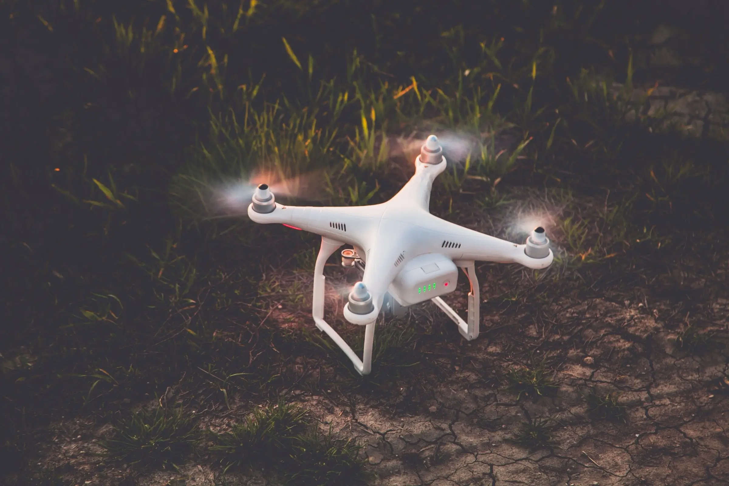 A white quadcopter with its propellers spinning, a moment from touching down on dry, cracked ground.